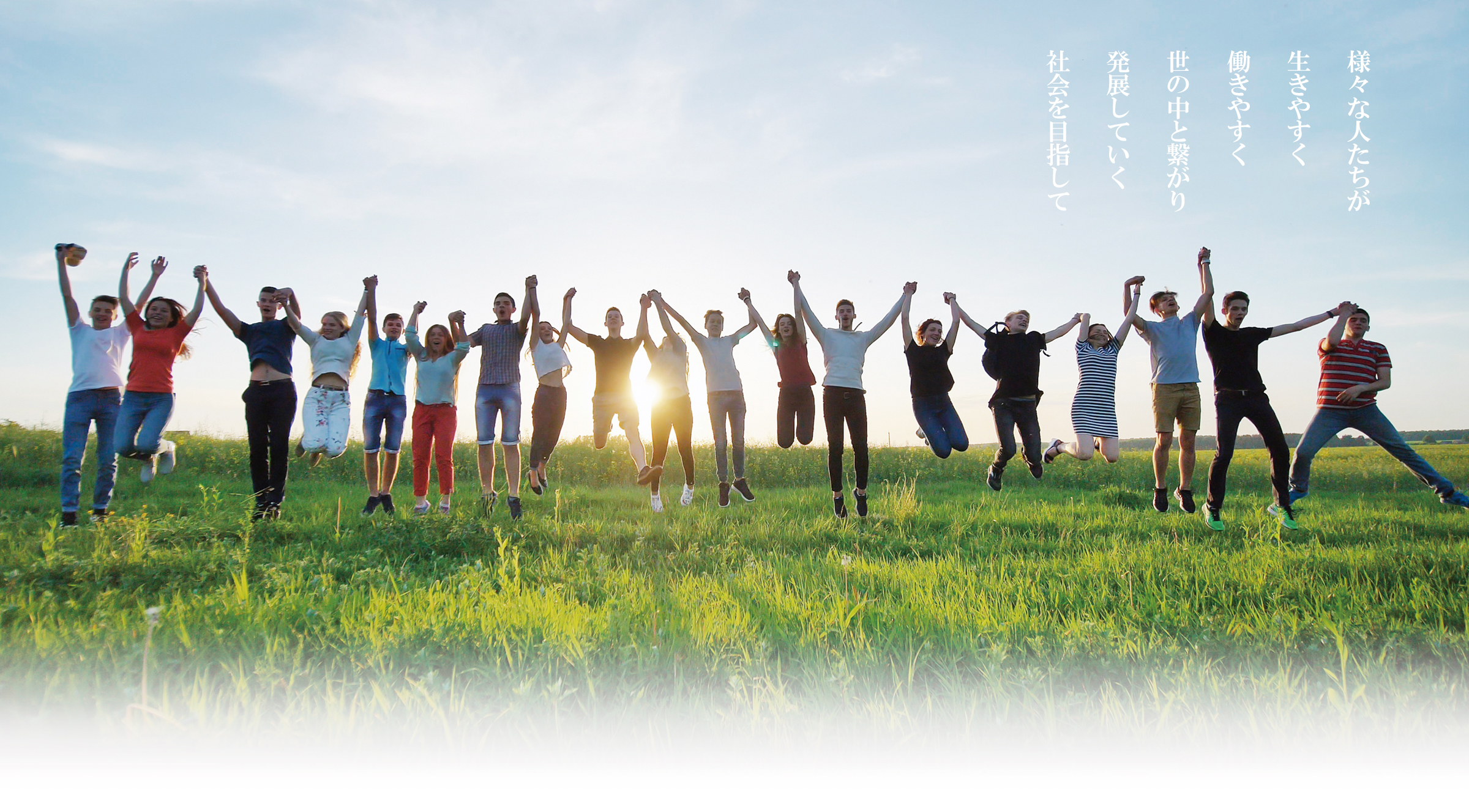 太陽を背に草原で一列になって笑顔で手をつないでいる19名の若者の写真